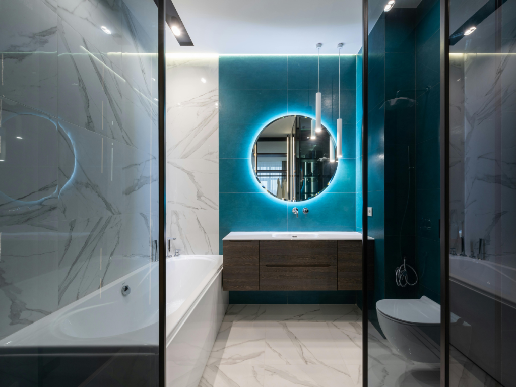 Erleben Sie ein modernes Badezimmer mit Spring Clearance-Atmosphäre, mit einem beleuchteten runden Spiegel, einer türkisfarbenen Akzentwand, Marmorfliesen, einer Badewanne und einem schwebenden Waschtischunterschrank.