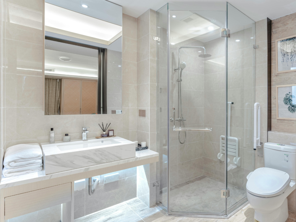 Modern badkamerontwerp met een glazen douche, wit toilet en wastafel met spiegel. Beige tegels bedekken elegant de muren en vloer, terwijl handdoeken en toiletartikelen netjes zijn gerangschikt voor het gemak.