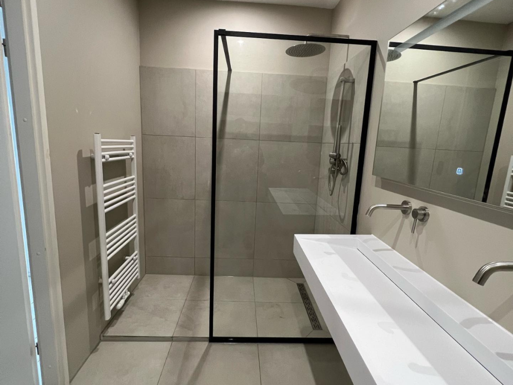 Das moderne Badezimmer verfügt über ein elegantes Innendesign mit einer Duschkabine aus Glas, einem Regenduschkopf, einem an der Wand montierten Handtuchheizkörper und einem Doppelwaschbecken mit verchromten Wasserhähnen. Elegante graue Fliesen bedecken nahtlos sowohl den Boden als auch die Wände.