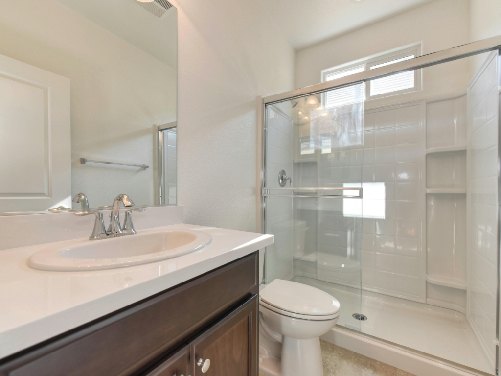 Ein modernes Badezimmer mit Badezimmerspiegeln über dem braunen Waschbecken mit einem Waschbecken mit verchromter Armatur. Genießen Sie die elegante Glasdusche, die weiße Toilette und ein weiß gefliestes Duschbad unter einem schmalen Fenster.