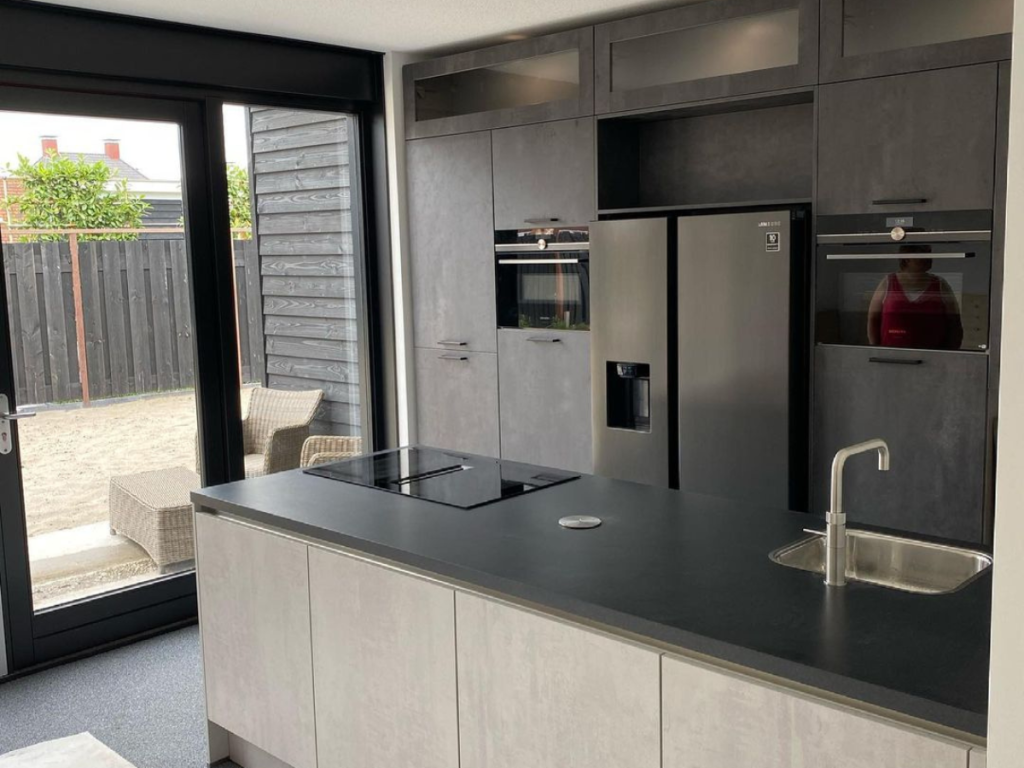 Moderne Küche mit dunklen Schränken, Einbaugeräten und einer Insel mit Spüle und Induktionskochfeld. Eine große Glastür öffnet sich zum Außenbereich mit einladenden Korbmöbeln und bietet einen nahtlosen Übergang, der direkt aus einer technischen Zeichnungsküche zu kommen scheint.