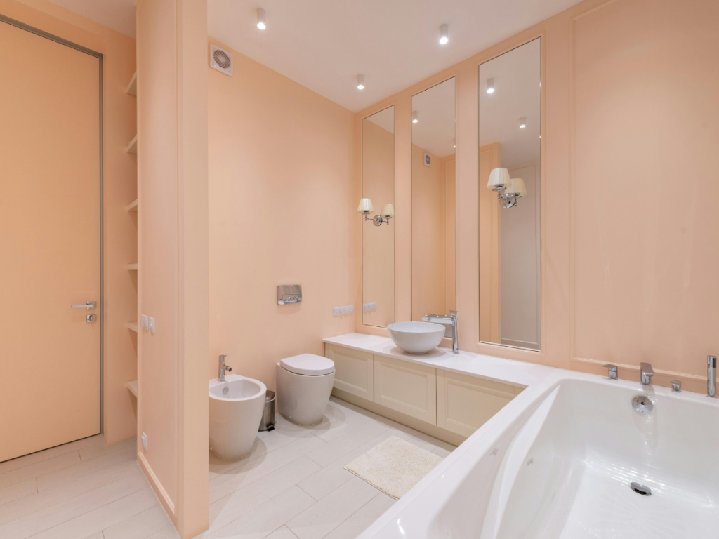 Geräumiges Badezimmer mit Badewanne, Waschbecken, großem Spiegel, WC und Bidet. Die Wände sind hell pfirsichfarben gestrichen und der Fußboden ist in einer beruhigenden hellen Farbe gefliest, alles harmonisch gestaltet, um sich nahtlos in das Rohrleitungsschema einzufügen.