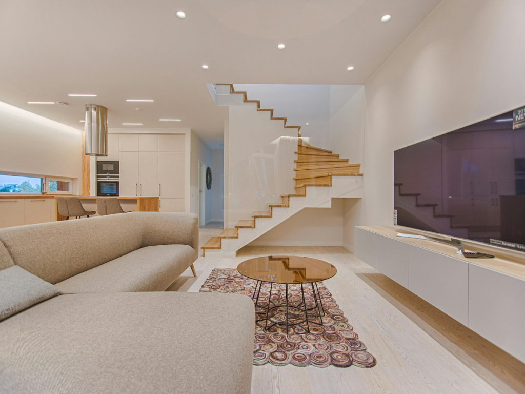 Ein modernes Wohnzimmer mit einem beigen Sofa, einem runden Couchtisch aus Glas, einem gemusterten Teppich und einem großen Flachbildfernseher. Die Holztreppe mit Glasgeländer ergänzt die natürlichen Elemente, während im Hintergrund der Küchenbereich sichtbar ist.