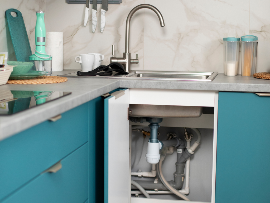 Die Küche ist mit Wasserleitungen unter der Spüle ausgestattet, die unter blaugrünen Schränken versteckt sind, ergänzt durch eine Arbeitsplatte mit verschiedenen Küchenutensilien und einen eleganten Edelstahlhahn.