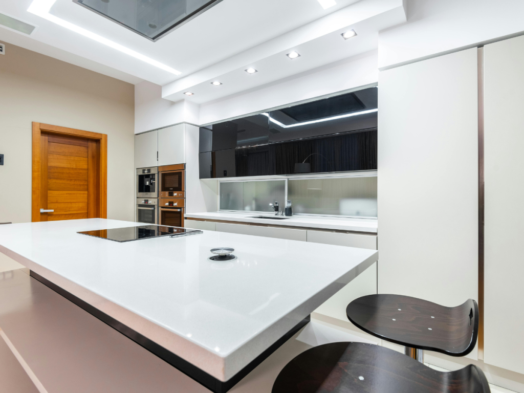Moderne Küche mit einer eleganten weißen Kochinsel, eingebautem Backofen, Mikrowelle, Induktionskochfeld und stilvollen schwarzen Barhockern. Eine helle Holztür und eine Einbaubeleuchtung vervollständigen dieses von Spring Clearance inspirierte Design.