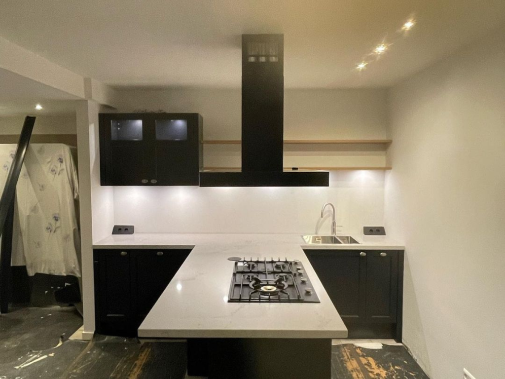 Moderne Küche mit einladender Atmosphäre, schwarzen Schränken, weißen Arbeitsplatten und einer zentralen Insel mit Herd. Minimalistisches Design mit Dunstabzugshaube, Waschbecken und Einbaubeleuchtung.