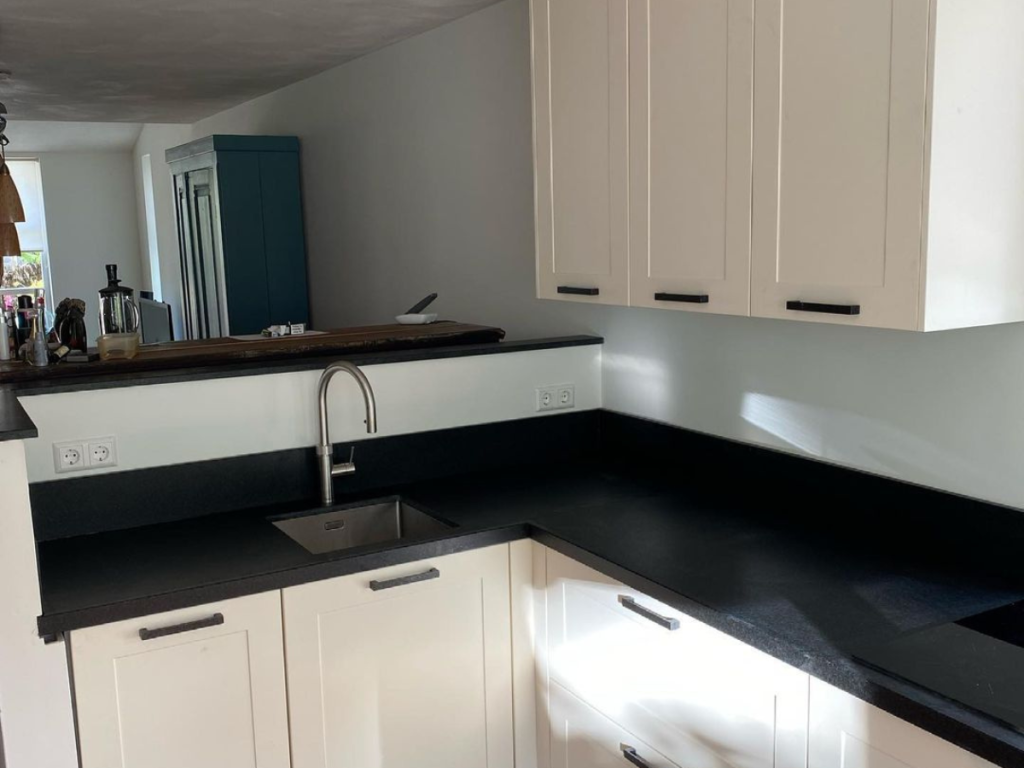 Moderne, einladende Küche mit weißen Schränken, schwarzen Arbeitsplatten und einer kleinen Spüle aus Edelstahl. Dunkle Schrankgriffe ergänzen das Design, während ein teilweise sichtbarer blau-grüner Schrank den Hintergrund mit Farbtupfer abhebt.