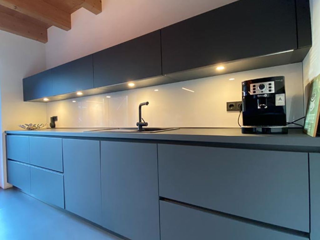 Moderne Küche mit eleganten, mattschwarzen Schränken und Arbeitsplatten, Unterschrankbeleuchtung, einer eingebauten Spüle und einer Kaffeemaschine auf der rechten Seite. Deckenbalken aus Holz sind sichtbar und bieten Frühlingstipps für alle, die einen anspruchsvollen und dennoch funktionalen Raum suchen.