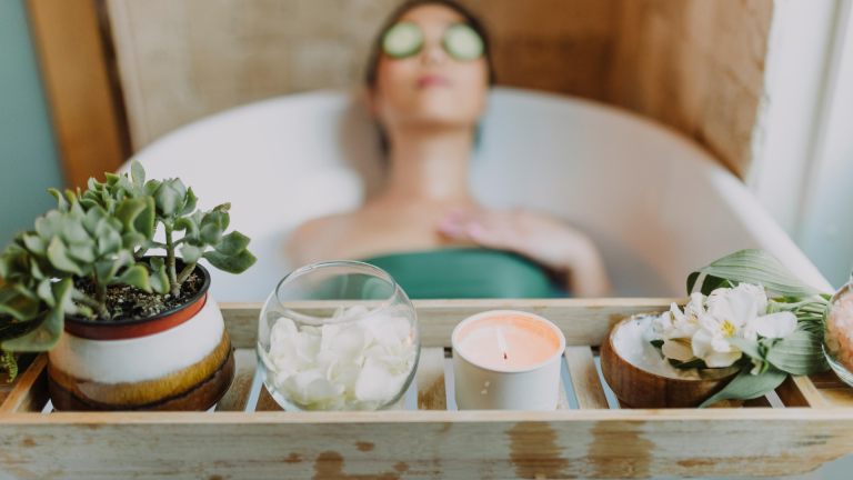 Eine Person entspannt sich in der Badewanne mit Gurkenscheiben auf den Augen, umgeben von Kerzen, Blumen und Frühlingspflanzen auf einem Holztablett.
