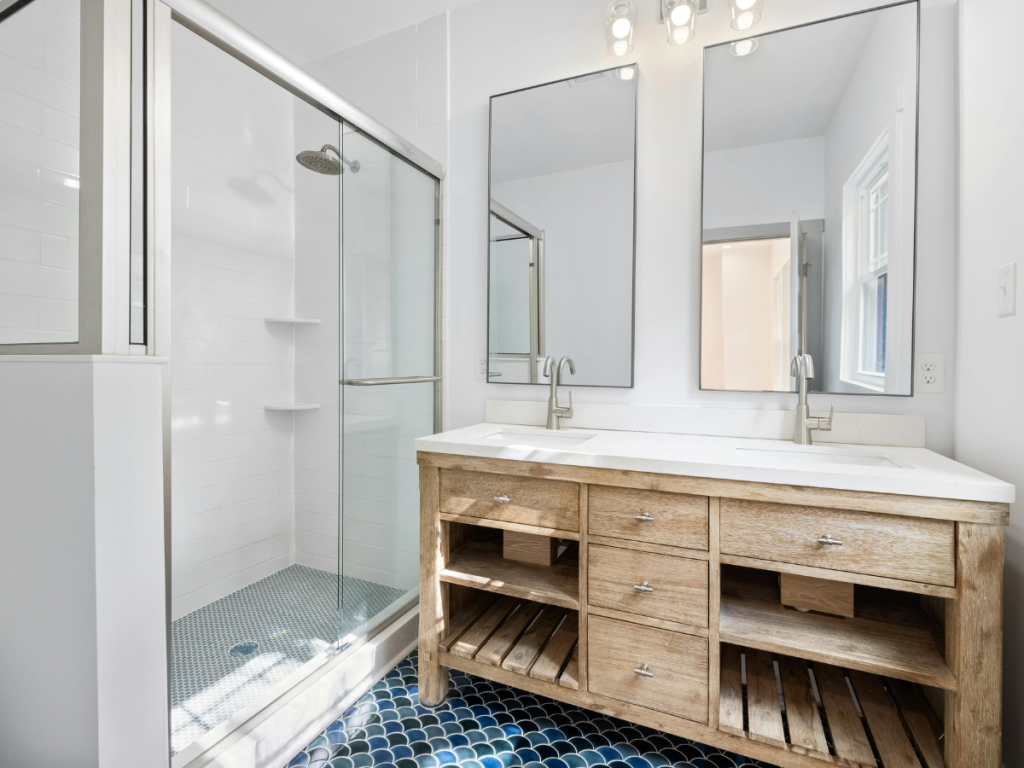 Das Badezimmer verfügt über ein Holzwaschbecken mit Doppelwaschbecken als Teil der kundenspezifischen Lösungen, ergänzt durch große Spiegel und eine Glasdusche. Blaue Fliesenböden und weiße Wände werden durch ein kleines Fenster durch natürliches Licht beleuchtet.