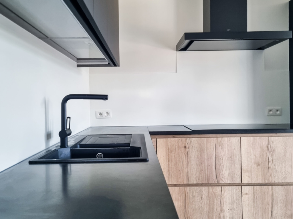 Moderne, einladende Küche mit einem schwarzen Wasserhahn über einem Waschbecken, ergänzt durch schwarze Arbeitsplatten und Holzschränke. Eine schlanke Dunstabzugshaube hebt sich von der makellosen weißen Wand ab und schafft eine einladende Atmosphäre.