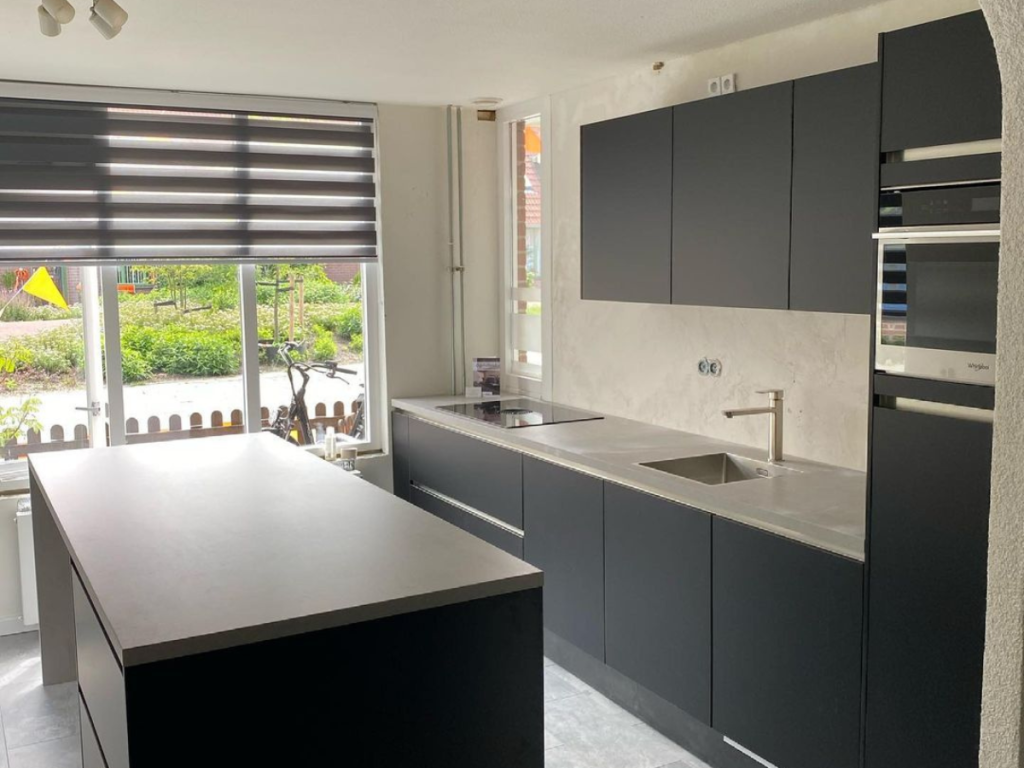 Moderne Küche in monochromer Farbpalette mit schwarzen Schränken, grauen Arbeitsplatten, einer Kücheninsel und einem Wasserhahn aus Edelstahl. Ein großes Fenster mit gestreiften Jalousien gibt den Blick in den Garten frei.