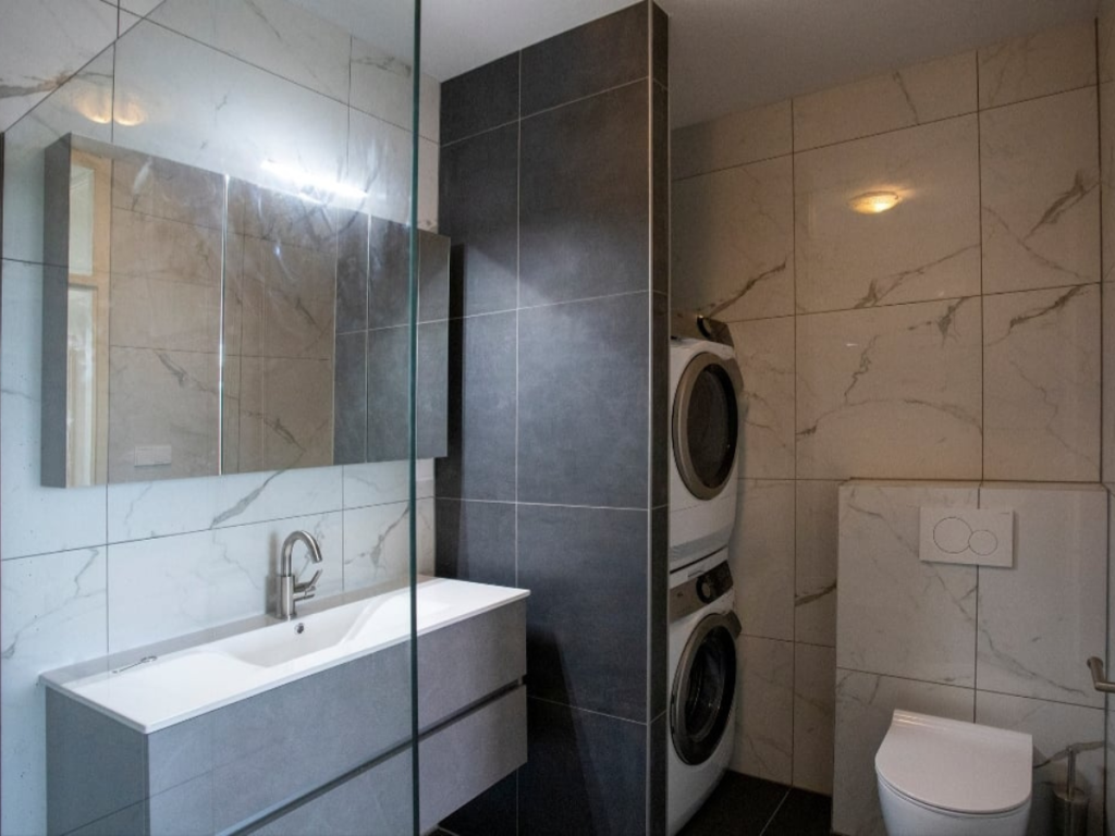 Modernes Baddesign mit grauen und weißen Fliesen, einem Wandwaschbecken mit Spiegel darüber, einer stapelbaren Waschmaschine und einem Trockner sowie einer Toilette.