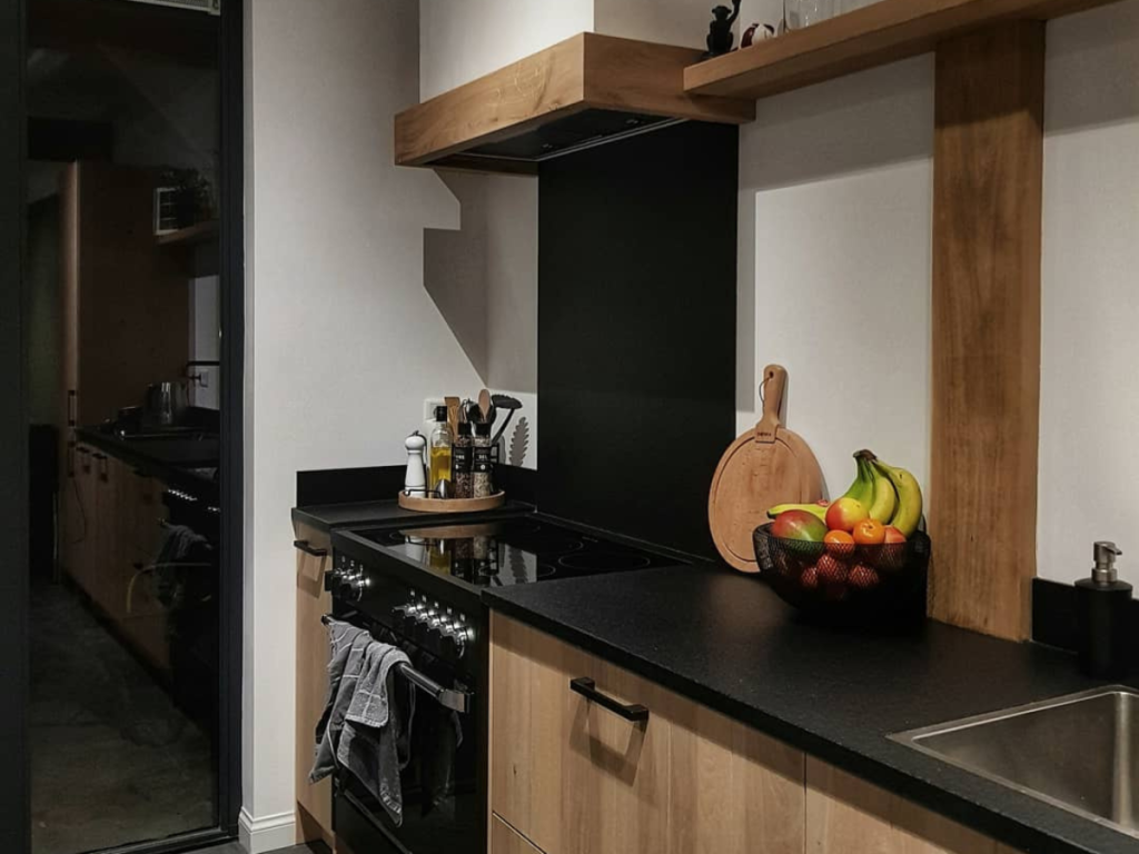 Moderne Küche mit schwarzen Arbeitsplatten, Holzschränken und stilvollen Regalen, die die neuesten Trends bei Küchengeräten präsentieren. Ein Obstkorb mit Bananen und Orangen steht auf der Theke neben einem Herd und einer Spüle.