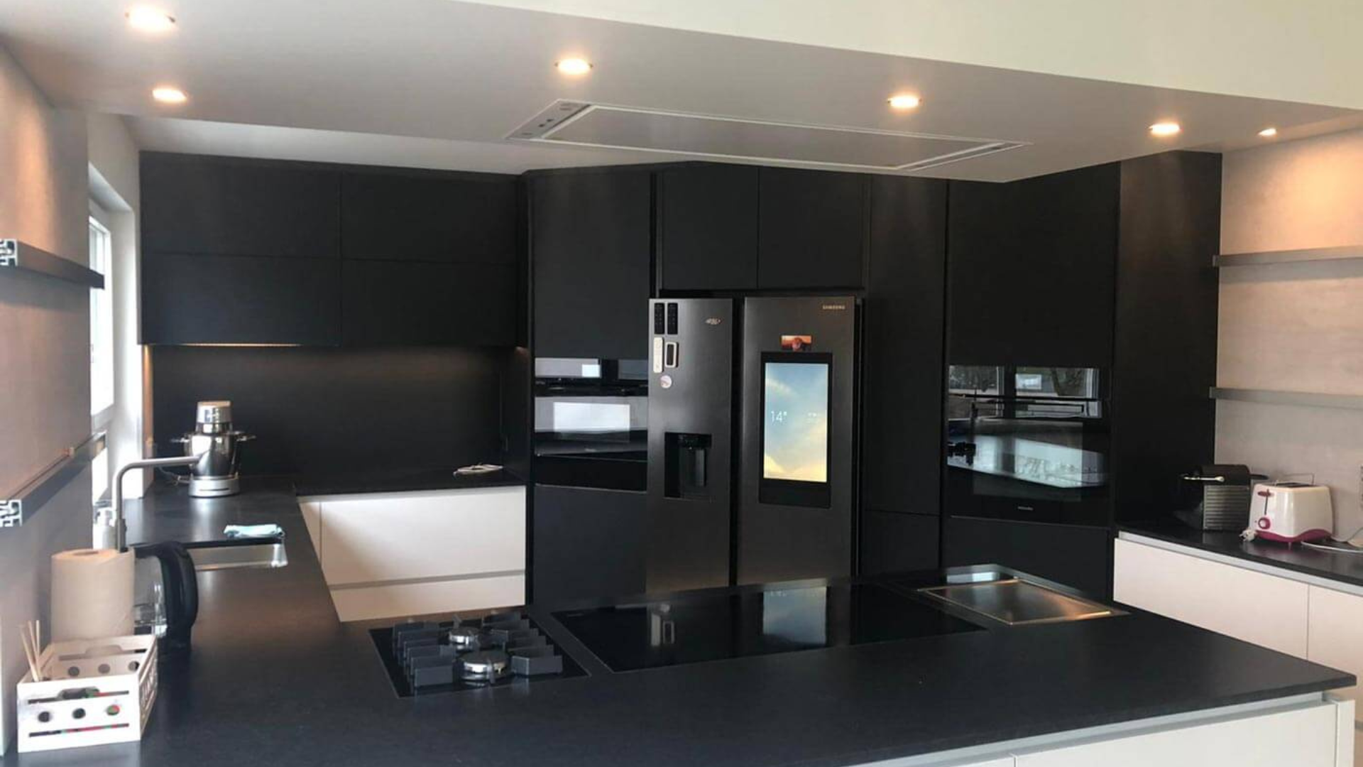 Eine moderne Küche mit einer monochromen Farbpalette, mit eleganten schwarzen Schränken, einem großen Kühlschrank, einem eingebauten Backofen und einem Induktionskochfeld auf der Insel. Es verfügt über Edelstahlakzente und eine Kaffeemaschine auf der Arbeitsplatte für den perfekten Morgenkaffee.