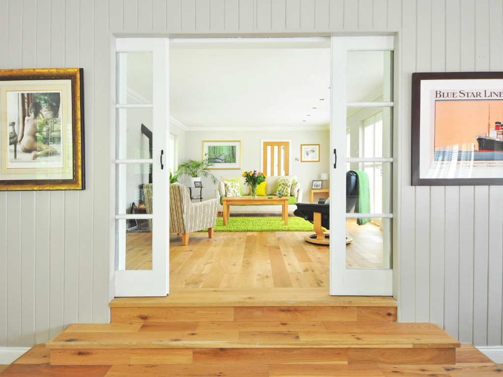 Ein Wohnzimmer mit Holzböden und Glasschiebetüren, die natürliche Elemente ins Innere bringen. Neutrale Wände ergänzen die Einrichtung, die aus gerahmter Kunst, einem grünen Teppich, Stühlen, einem Couchtisch und einer Blumenvase besteht.
