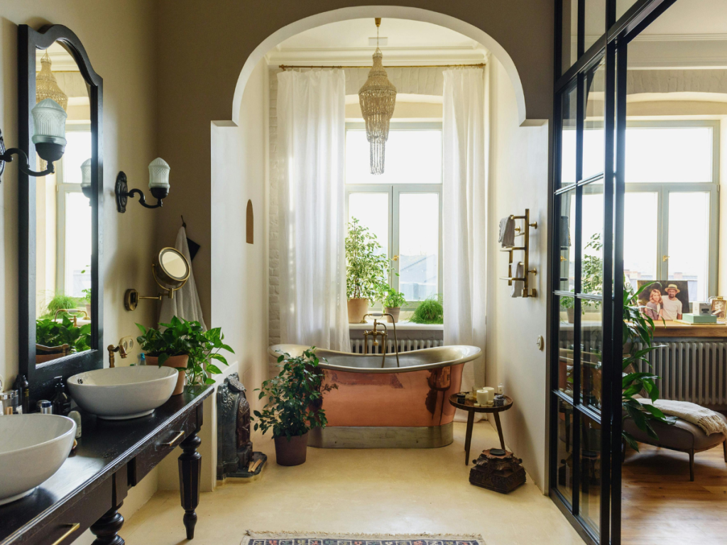 Een elegante badkamer met een vrijstaand bad, grote ramen en planten die de ruimte tot leven brengen. Dubbele wastafel met ronde wasbakken en een wandspiegel versterken de charme. Warme verlichting maakt de gezellige sfeer compleet.