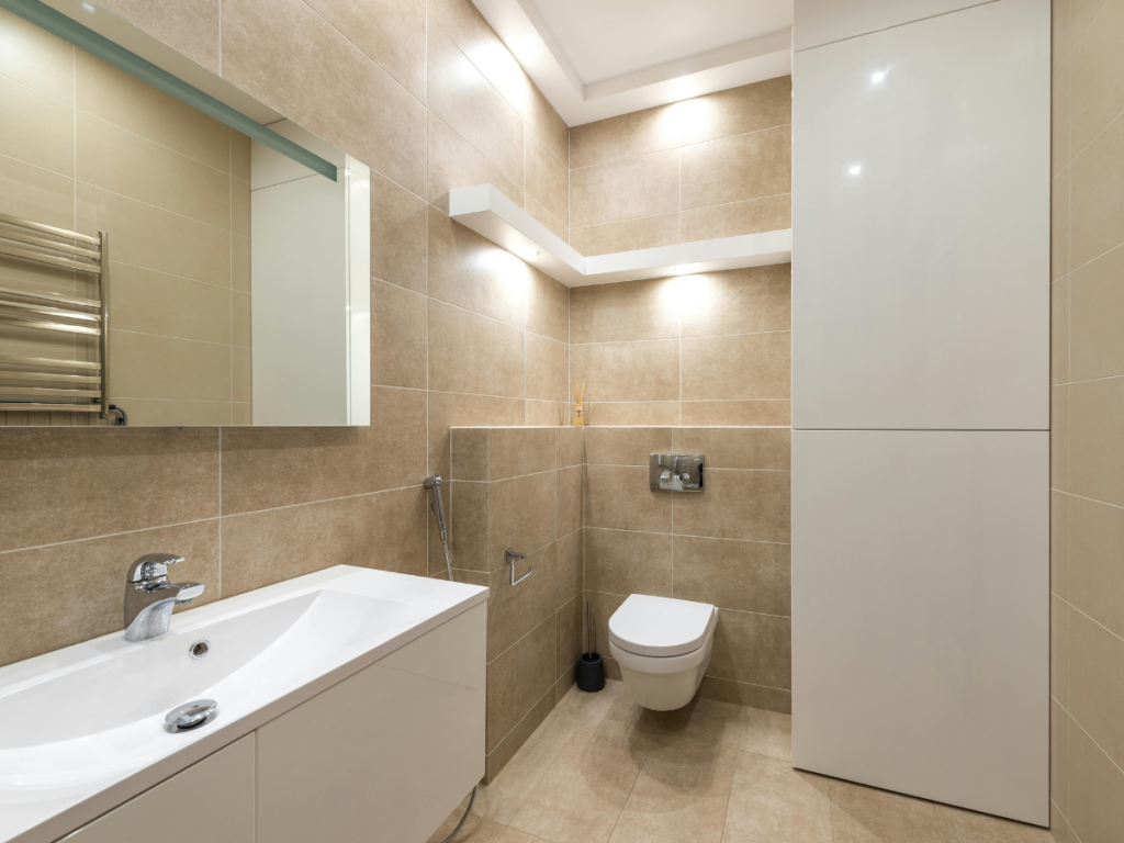 Modernes Badezimmer mit beigen Fliesen, einem Wandschrank, Waschbecken mit Wasserhahn und Badezimmerspiegeln über dem großen Spiegel. Es verfügt über einen Handtuchhalter und einen weißen Aufbewahrungsschrank für einen sauberen und organisierten Look.