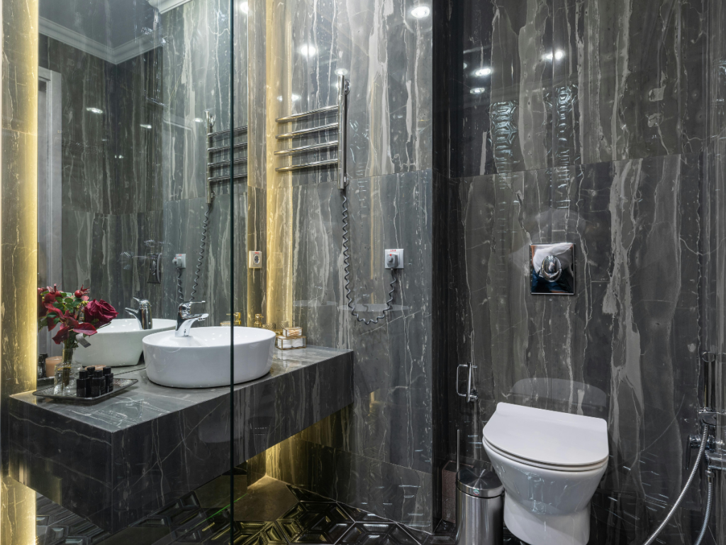 Een moderne badkamer heeft donkere marmeren muren, een waterbesparende wandwastafel en een toilet. Een spiegel, handdoekrek en een vaas met rode bloemen verfraaien het aanrecht.