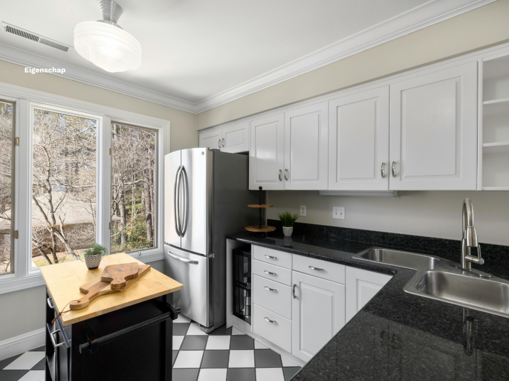Eine moderne Küche mit kariertem Fliesenboden, weißen Schränken, schwarzen Arbeitsplatten, einem eleganten Edelstahlkühlschrank, einer Kücheninsel aus Holz, Schneidebrettern und großen Fenstern mit Blick auf die Bäume.