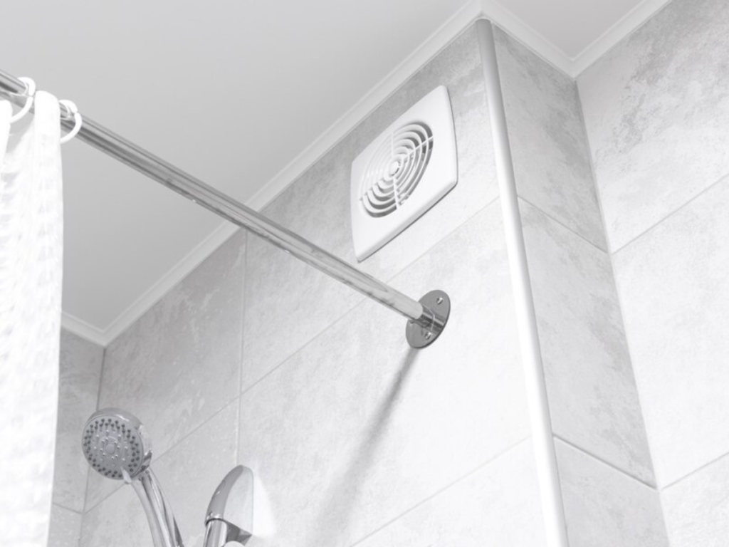 Badezimmerecke mit weißen Fliesen, einem montierten Duschkopf, einer Duschvorhangstange und einem Ventilator an der Wand.