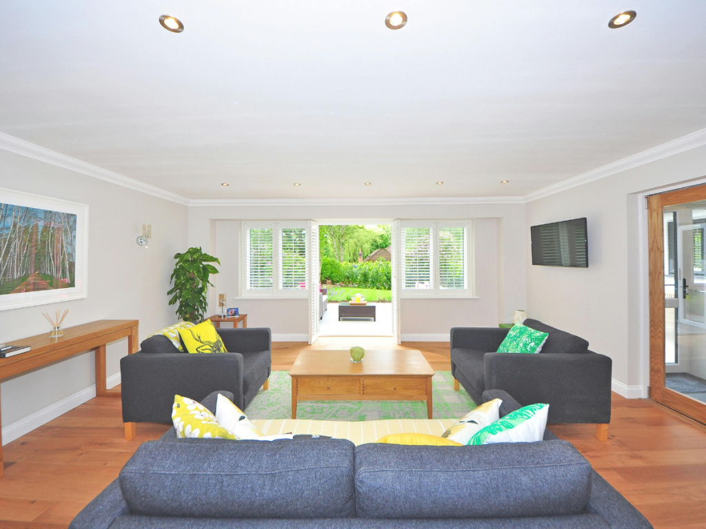 Ein geräumiges Wohnzimmer mit grauen Sofas, einem Couchtisch aus Holz und einem Fernseher an der Wand. Große Fenster lassen natürliches Licht herein und eine Tür öffnet sich zum Garten, so dass sich natürliche Elemente nahtlos in Ihr Zuhause einfügen.