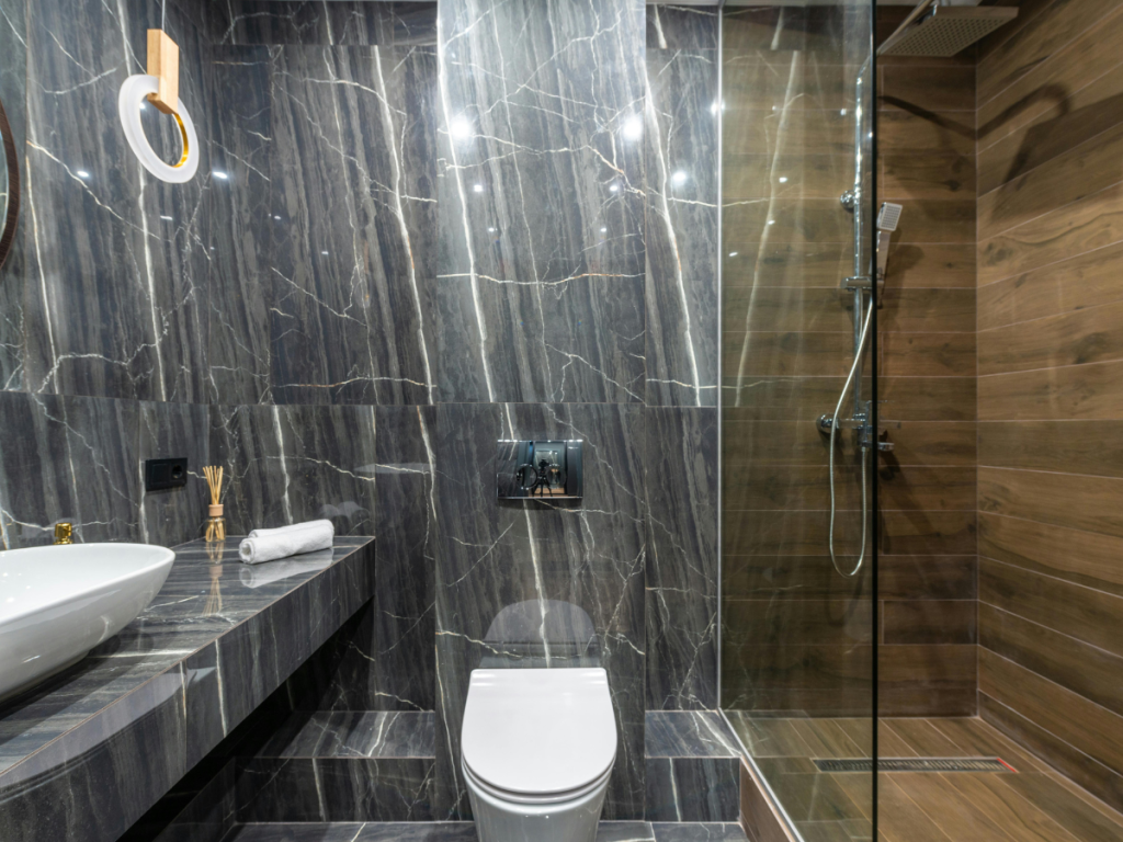 Moderne badkamer met donkere marmeren tegels, een wit wandcloset en een waterbesparende glazen douchecabine. Het beschikt over een wastafel, opgerolde handdoek en een ronde spiegel met licht.