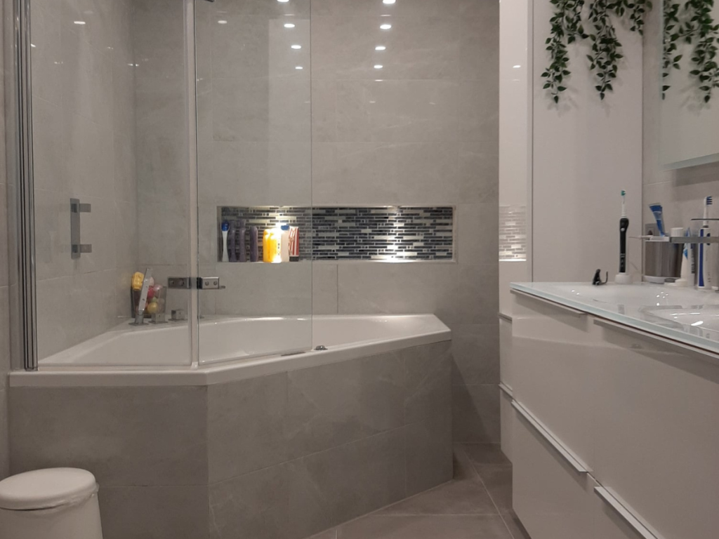 Moderne badkamer met glazen hoekbad, dubbele wastafels op een wit wastafelmeubel en aan de muur gemonteerde toiletartikelen. Grijze tegelwanden en vloer; een groene plant voegt charme toe aan de onlangs voltooide badkamerrenovatie.
