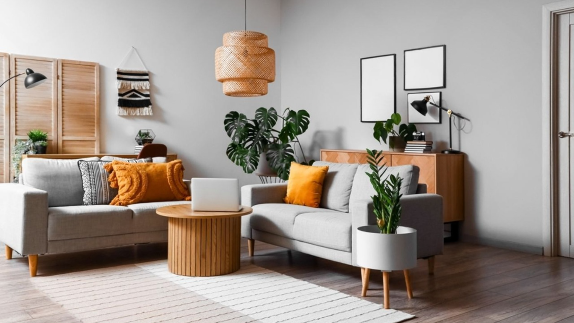 Modernes Wohnzimmer mit grauen Sofas, orangefarbenen Kissen und einem runden Couchtisch aus Holz, der mit natürlichen Elementen dekoriert ist. Verschiedene Pflanzen bringen Leben in den Raum. Zwei gerahmte Fotos hängen an der Wand unter einer geflochtenen Hängelampe darüber.