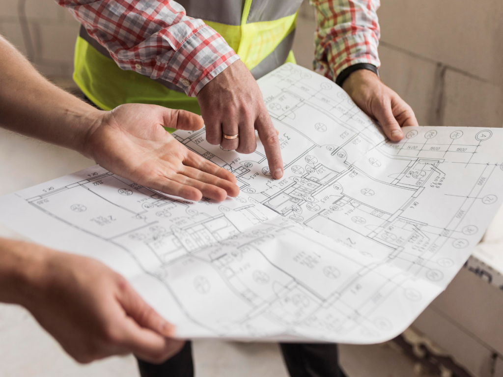 Zwei Personen schauen sich einen detaillierten architektonischen Entwurf an und zeigen auf hervorgehobene Bereiche für Küchen und Bäder in einer belebten Bauumgebung.