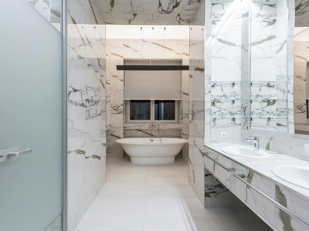 Een moderne badkamer met marmeren muren en vloer, een vrijstaand bad, een wastafel met dubbele wastafel en een grote spiegel. Ook zijn er waterbesparende voorzieningen om de duurzaamheid te vergroten.