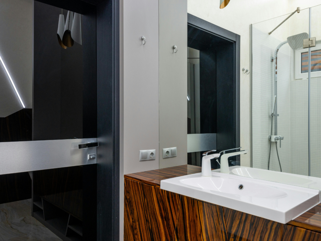 Modernes Badezimmer mit Waschbecken mit Holzmotiv, weißem Waschbecken, Seifenspender an der Wand, Spiegel, Dusche mit Glastür und Wänden in neutralen Tönen.