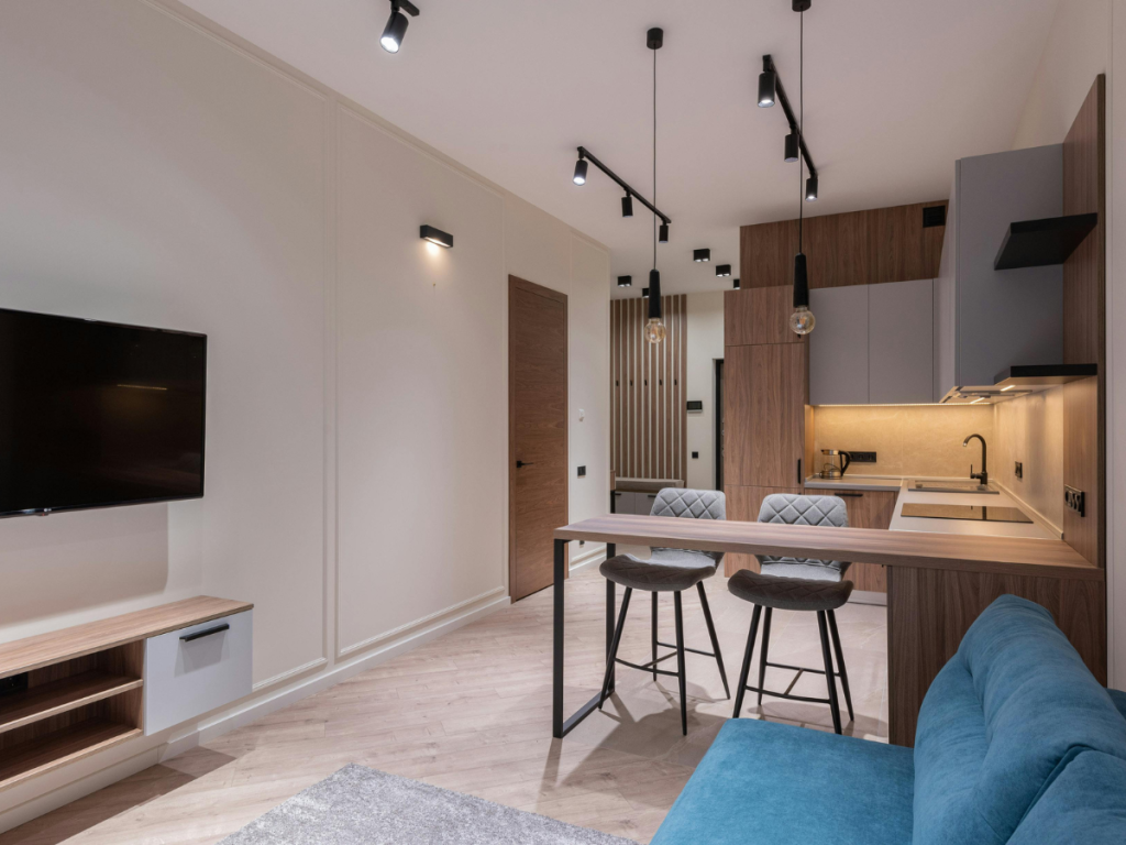 Modernes Studio-Apartment mit Küche und Wohnzimmer, Essbereich, montiertem TV und blauem Sofa, mit Holzakzenten und minimalistischer Beleuchtung.