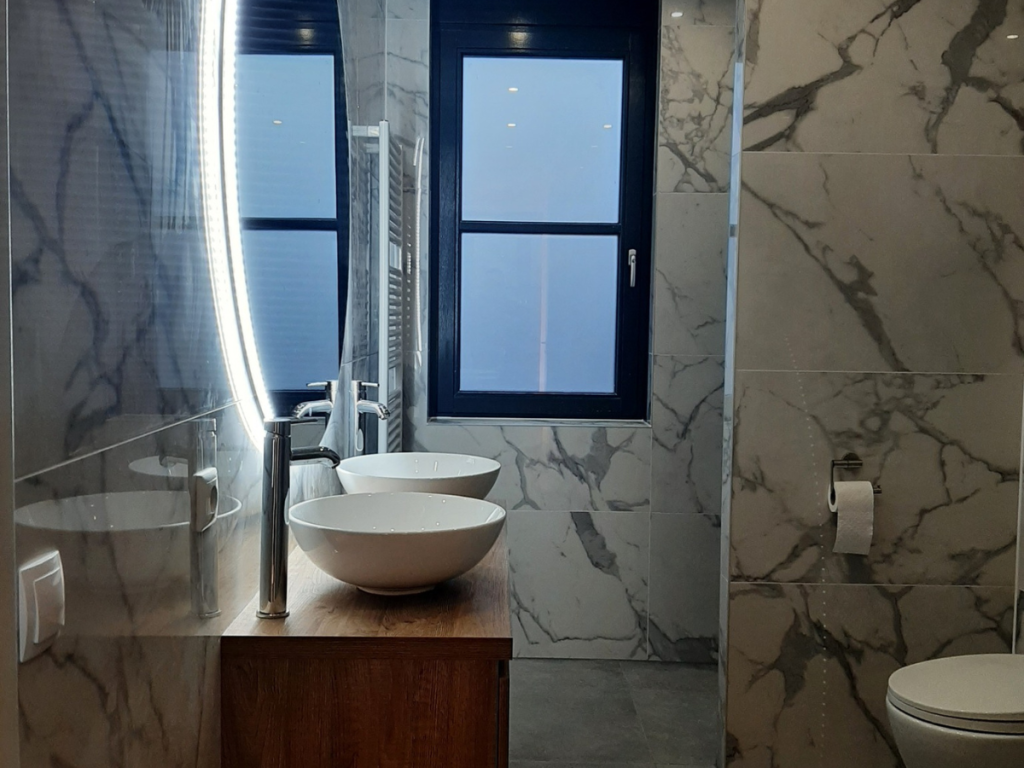 Modernes Badezimmer mit Marmorwänden, einem großen beleuchteten Spiegel, zwei Aufsatzwaschbecken auf einer Holzplatte, dezenten Farbakzenten, einem Fenster mit schwarzem Rahmen und einer Toilette mit wandmontiertem WC-Rollenhalter.