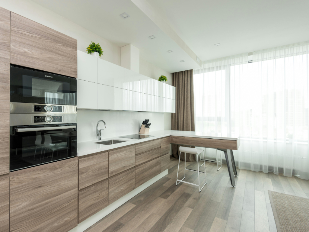 Moderne Küche mit hellen Holzschränken, weißen Arbeitsplatten, eingebautem Backofen, Induktionskochfeld und einem minimalistischen Essbereich mit einem Hocker an großen Fenstern mit transparenten Vorhängen.