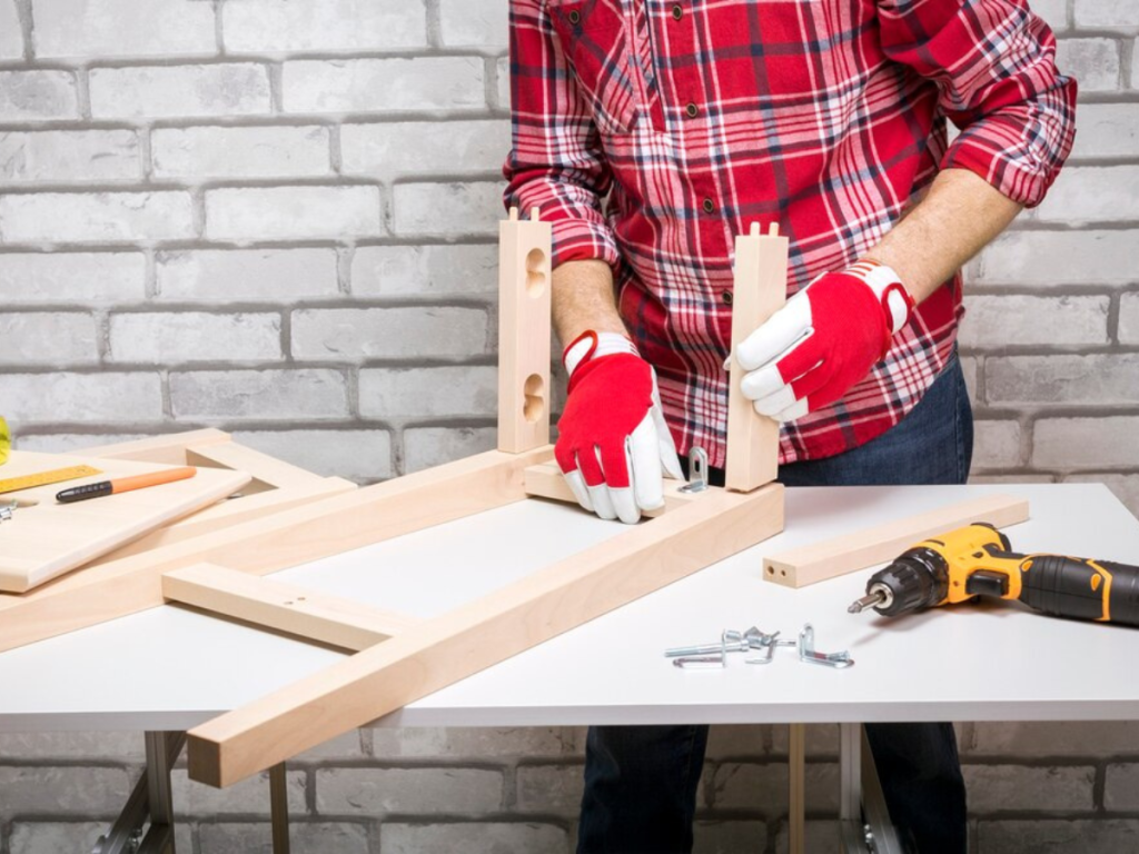 Eine Person trägt rote Handschuhe und ein kariertes Hemd und baut Holzmöbel auf einem Tisch zusammen. Dafür verwendet er Schrauben und einen Bohrer. Die Möbel stehen an einer weißen Backsteinwand.
