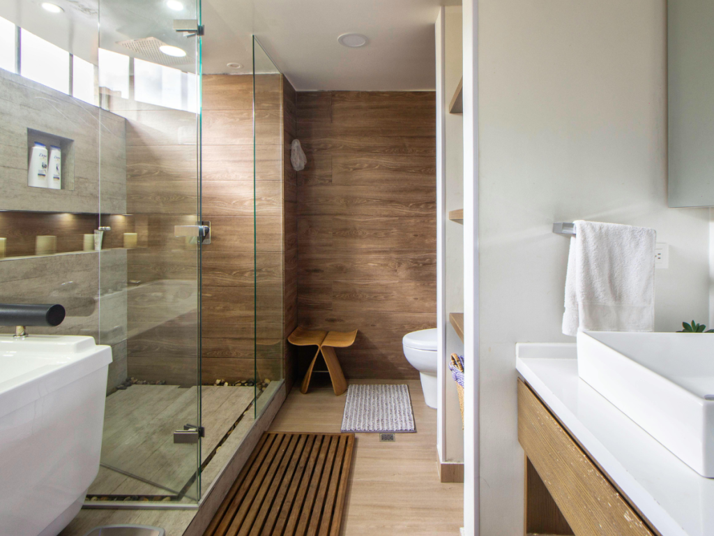 Modernes Badezimmer mit Holzakzenten, ausgestattet mit einer Duschkabine aus Glas, weißem Waschbecken, WC, Holzhocker, Handtuchhalter und großem Spiegel. Kundenfeedback hebt das natürliche Licht hervor, das durch die hohen Fenster hereinströmt.