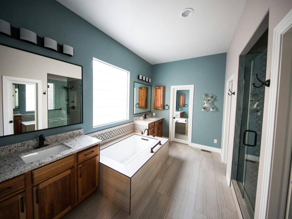 Modernes Badezimmer mit Holzschränken, einer großen Badewanne, einer Glasdusche, zwei Waschbecken, Wandspiegeln und blaugrünen Wänden mit neutralem Fliesenboden.