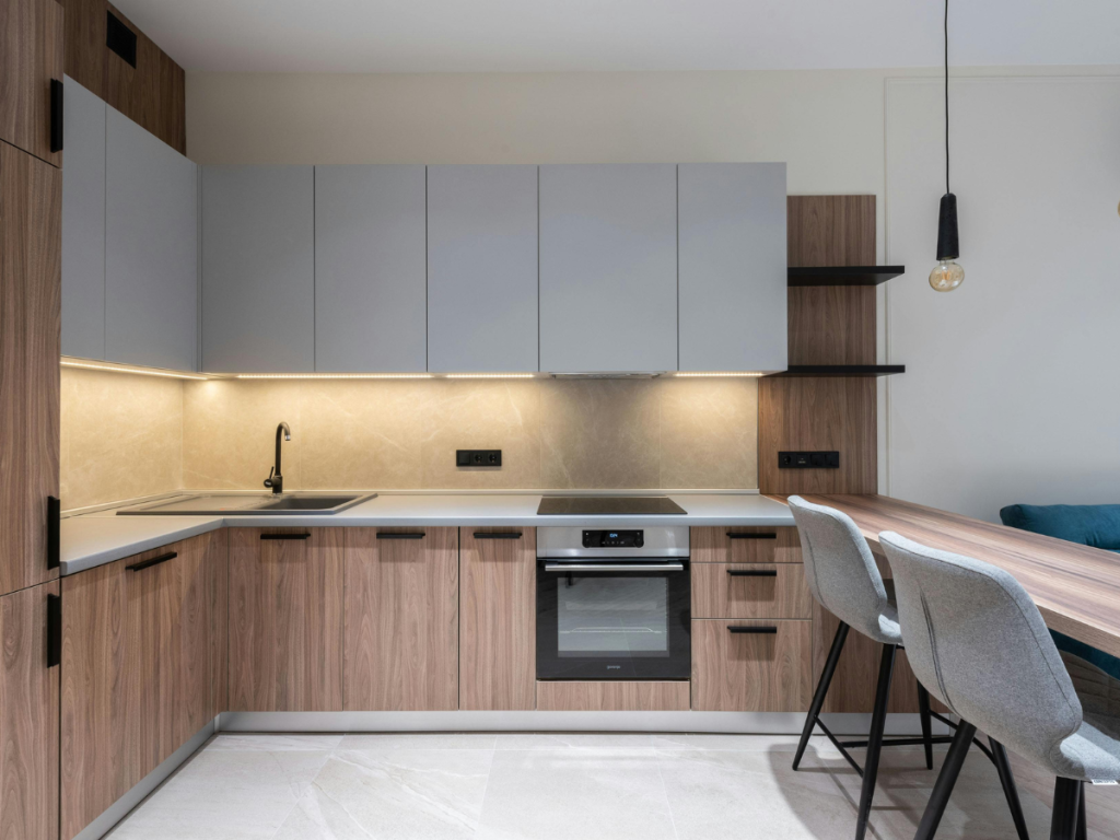 Moderne Küche mit Unterschränken aus Holz, grauen Schrankwänden, Einbaubackofen, Elektroherd, Spüle, zwei grauen Barhockern und einer Pendelleuchte über der Frühstückstheke. Alle mit einem schlanken, offenen Regaldesign für einen zeitgemäßen Look.