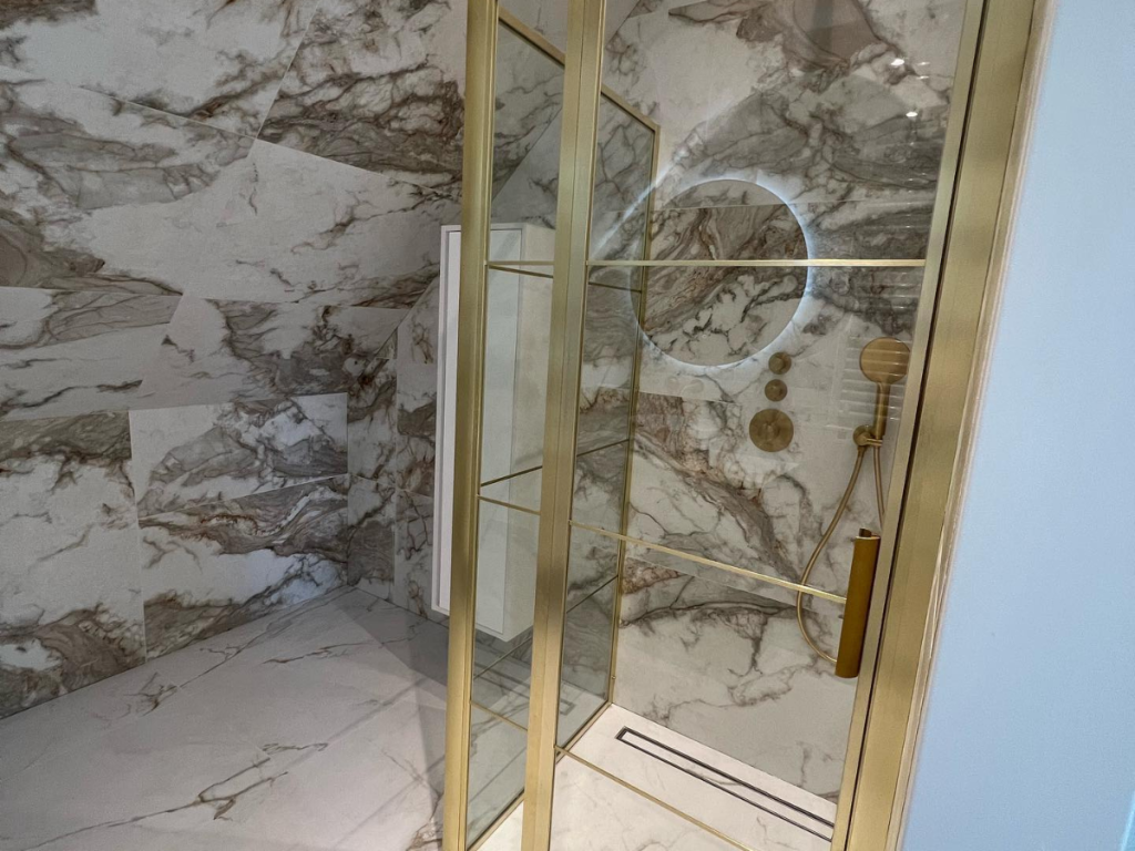 Die moderne Dusche mit goldenen Armaturen, Glastüren und Marmorwänden und -böden sorgt für luxuriöse Farben. Ein runder Spiegel mit Hintergrundbeleuchtung ist in der Dusche angebracht und unterstreicht sowohl den Stil als auch die Funktionalität.