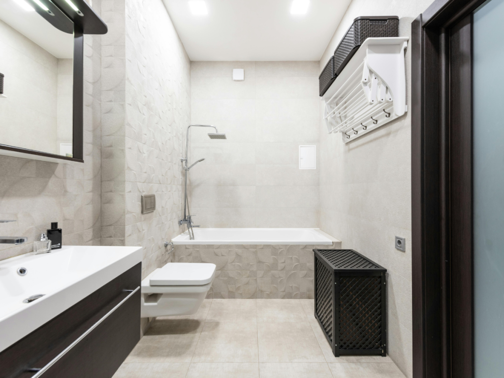 Modernes Badezimmer mit Badewanne, hängender Toilette, Waschbecken, Aufbewahrungskörben und neutralen Fliesenböden und -wänden, die durch eine stilvolle Beleuchtung für eine warme und einladende Atmosphäre aufgewertet werden.