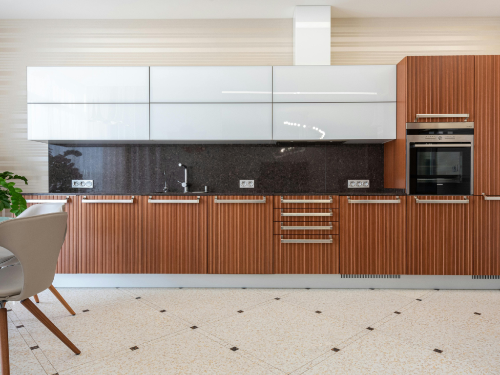 Moderne Küche mit Holz- und weißen Schränken, eingebautem Backofen, schwarzer Arbeitsplatte, Spüle und gemustertem Fliesenboden. Auf der linken Seite ist ein Teil eines beigen Stuhls zu sehen – perfekt für alle, die eine moderne Küche mit Stil und Funktionalität suchen.