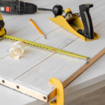 Maßband, Hobel, Bleistift und Bohrer liegen auf einem Holzbrett, das an einen Tisch geklemmt ist, daneben Holzspäne: das perfekte Werkzeug für Ihre nächsten DIY-Projekte.