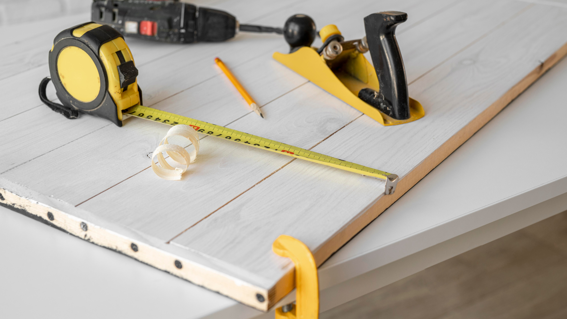 Maßband, Hobel, Bleistift und Bohrer liegen auf einem Holzbrett, das an einen Tisch geklemmt ist, daneben Holzspäne: das perfekte Werkzeug für Ihre nächsten DIY-Projekte.