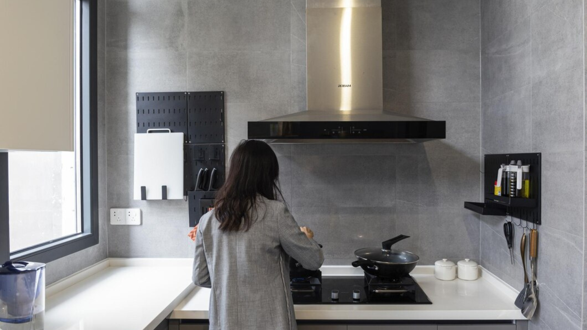 Eine Frau steht beim Kochen an einem modernen Herd in der Küche, umgeben von grauen Wänden, einer Dunstabzugshaube und ordentlich angeordneten Küchenutensilien an der Wand.