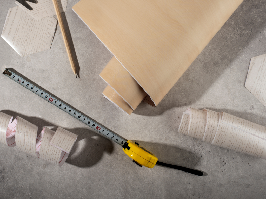 Ein Maßband, ein Bleistift, rechteckige Furnierblätter, sechseckige Fliesen und Tapetenrollen sind fein säuberlich auf einer grauen Fläche angeordnet: die perfekte Inspiration für Ihre nächsten DIY-Projekte.