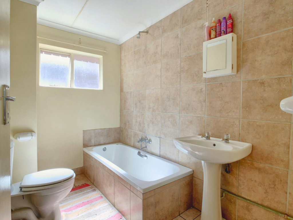 Ein Badezimmer mit Badewanne, WC, Waschbecken, Wandschrank, gefliesten Wänden, einem gestreiften Teppich in sanften Farbpaletten und drei Flaschen auf dem Schrank unter einem Fenster.