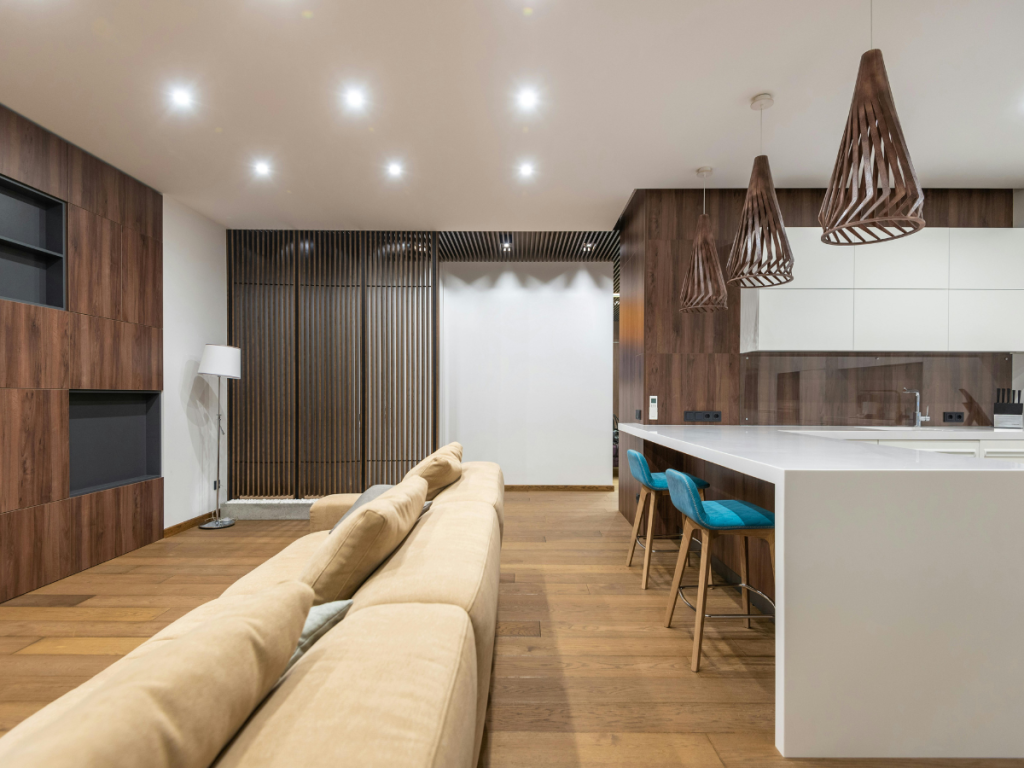 Moderne offene Küche und Wohnzimmer mit Holzakzenten, einem beigen Sofa, einer weißen Kücheninsel, türkisfarbenen Barhockern, Pendelleuchten und Einbaustrahlern.