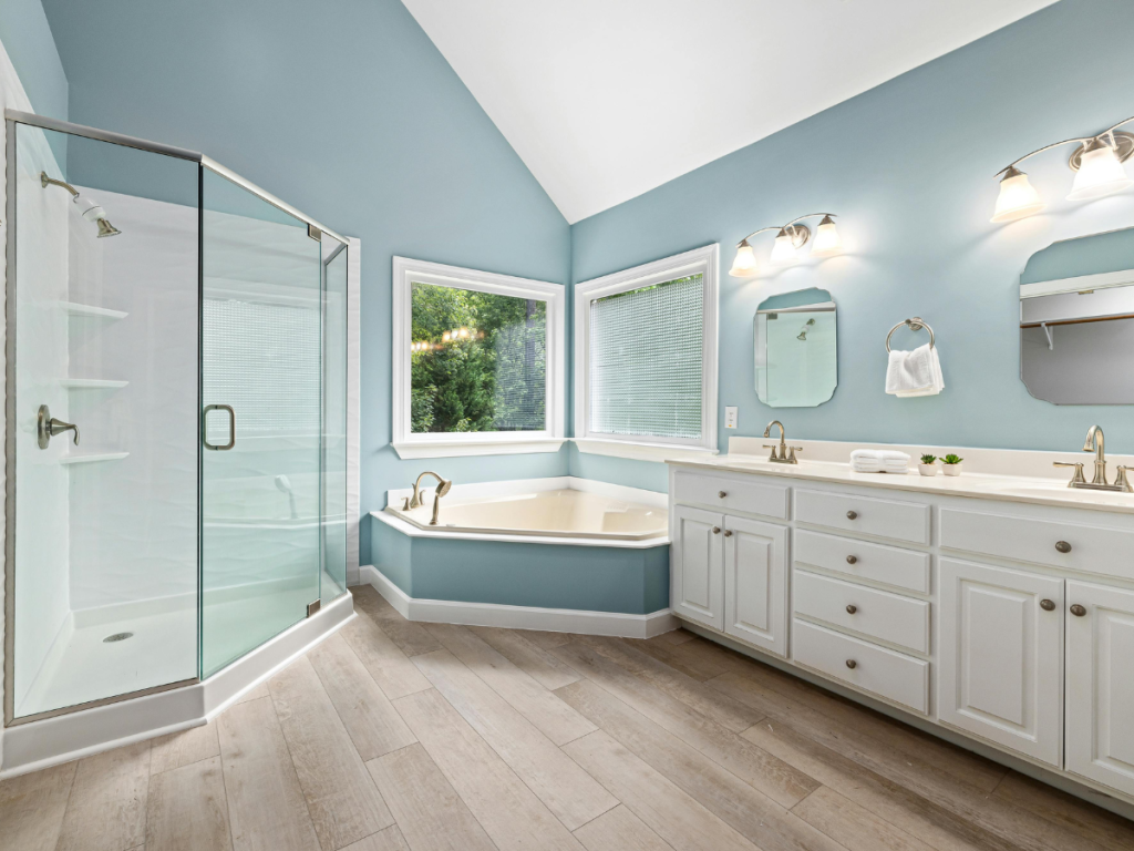 Modernes Badezimmer mit blauen Wänden in trendigen Farbpaletten, Glasdusche, Eckbadewanne, Doppelwaschbecken mit weißen Schränken, großen Fenstern und Fliesenboden in Holzoptik.