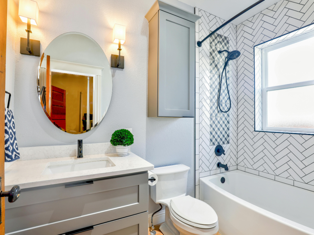 Modernes Badezimmer im eklektischen Stil, ausgestattet mit einem runden Spiegel, Wandarmaturen, grauem Waschbecken, WC, Badewanne mit Fischgrätmuster und einem Fenster.